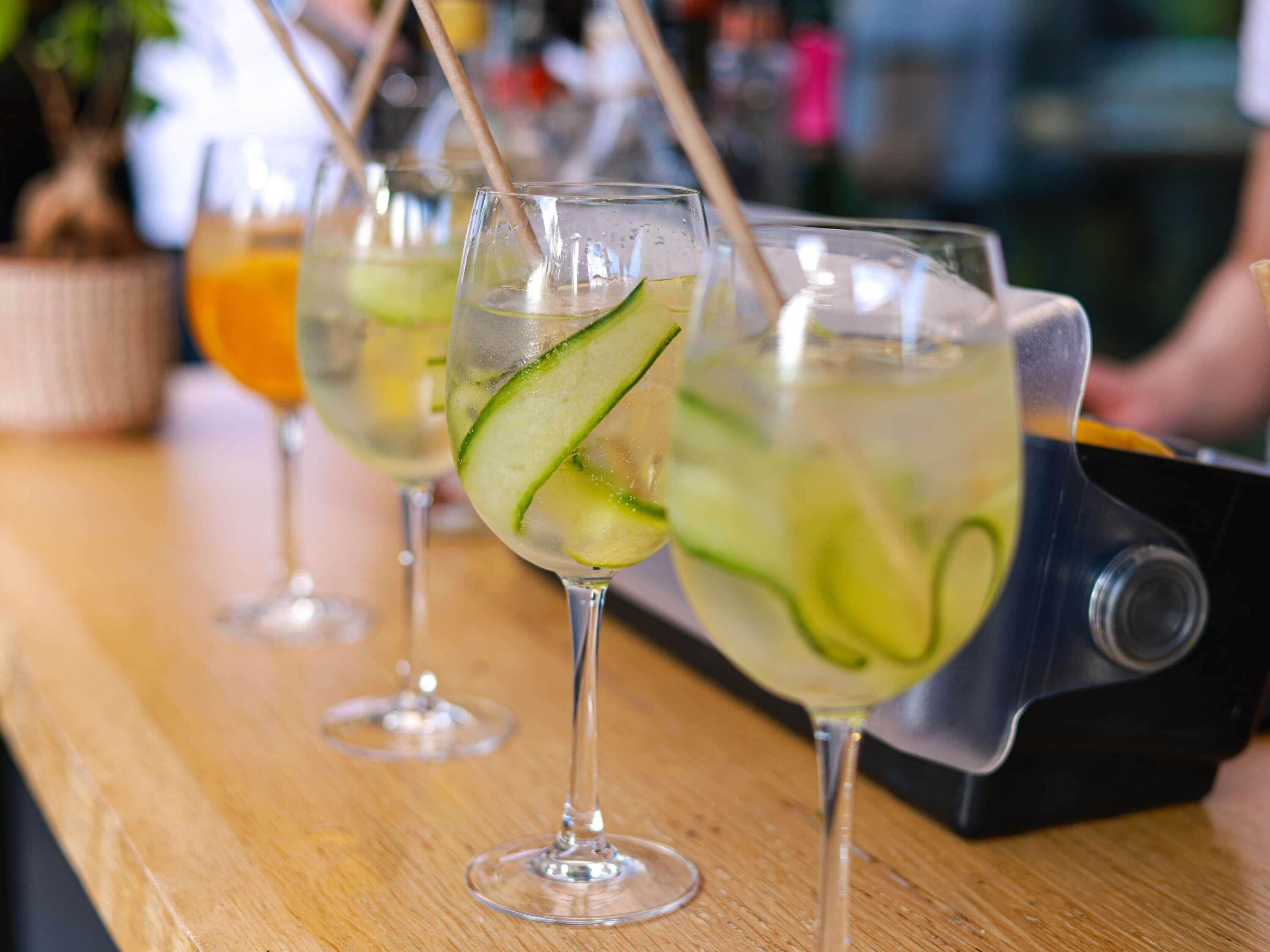 Tres vasos de una bebida con rodajas de pepino, sobre una barra de madera.