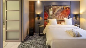 Hotelkamer met een bed, twee wandlampen, een groot abstract schilderij en een glazen deur naar een badkamer.