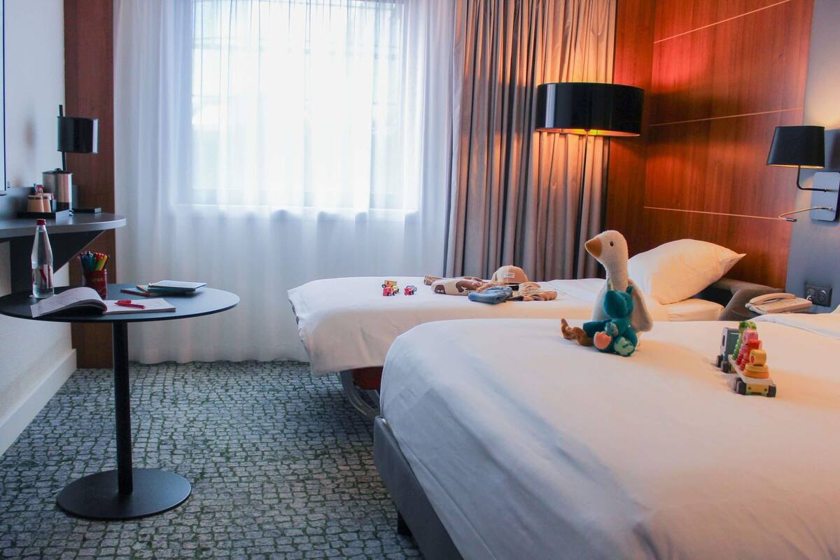 Une chambre d’hôtel avec deux lits, une petite table, une lampe et une fenêtre avec des rideaux.