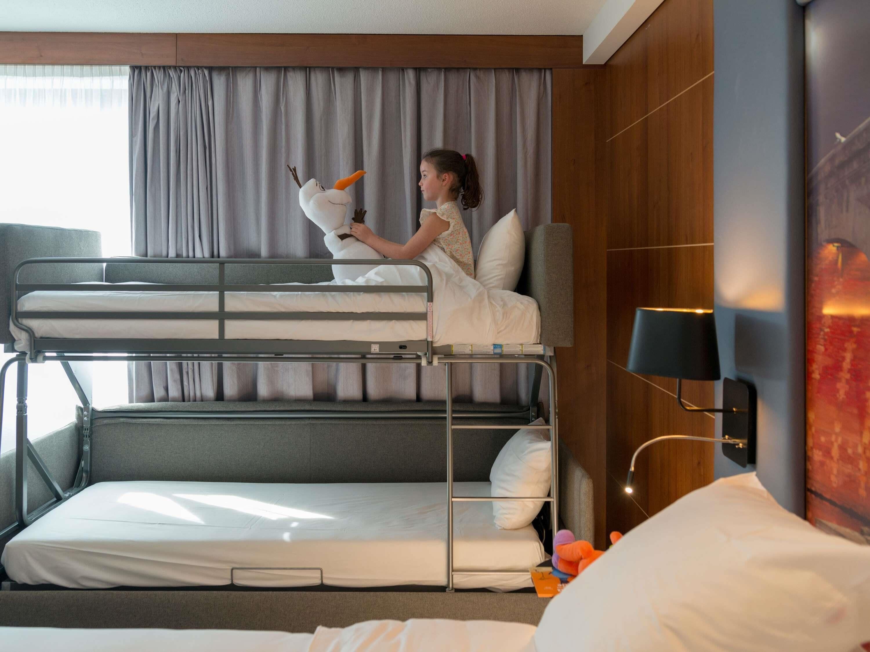 A bunk bed room with a child on the top bunk, a lamp, and a bed with toys.