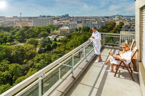 Un balcone con ringhiera in vetro che si affaccia su una città con spazi verdi ed edifici.