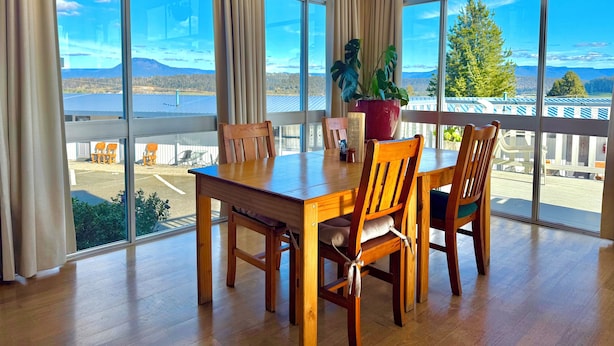 Uma área de refeições com mesa e cadeiras de madeira, janelas grandes com cortinas, um vaso de planta e vista para um cais e montanhas.