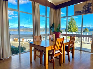 A dining area with a wooden table and chairs, large windows with curtains, a potted plant, and a view of a dock and mountains. - Mountain View Country Inn (Deloraine)