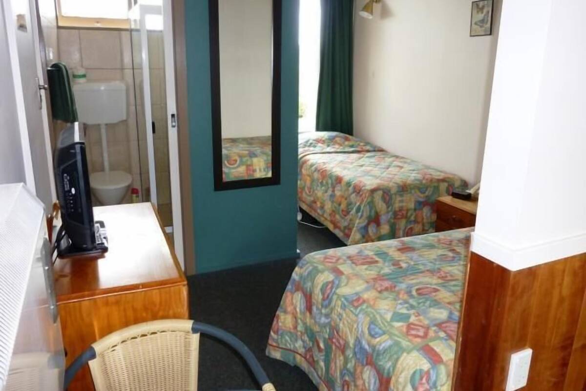A compact hotel room with two single beds, a small desk with a heater, a chair, a mirror, and a window with curtains.