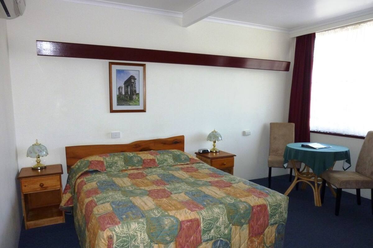 A hotel room with a bed, two bedside tables with lamps, a chair, a small table, and a window with curtains.