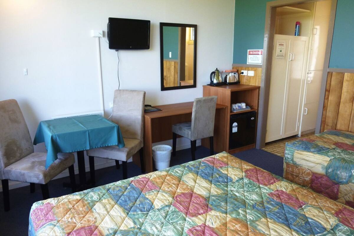 A motel room with a desk, two chairs, a TV, a small refrigerator, a cabinet, and two beds with colorful quilts.