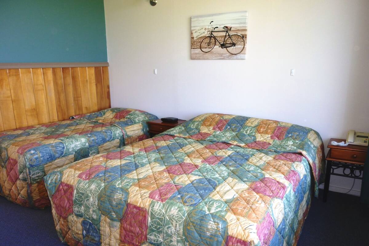 A room with two beds, each covered with a colorful quilt. There is a small table with a telephone on it, and a wall-mounted light fixture above the beds.