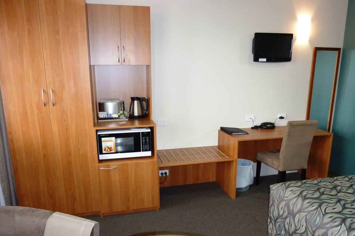 A compact room with a wooden wardrobe, a microwave, a small desk with a chair, a television, and a patterned bedspread.