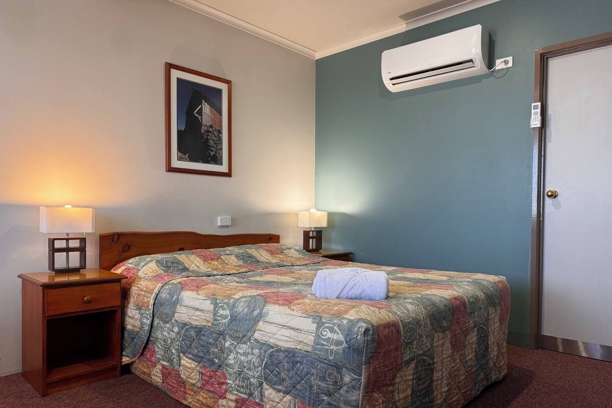 A hotel room with a bed, two bedside tables with lamps, an air conditioning unit, a door, and a framed picture on the wall.