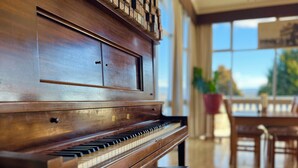 Um piano de cauda imponente numa sala ampla, com grandes janelas, chão de madeira e um espaço de refeições com cadeiras e mesa.