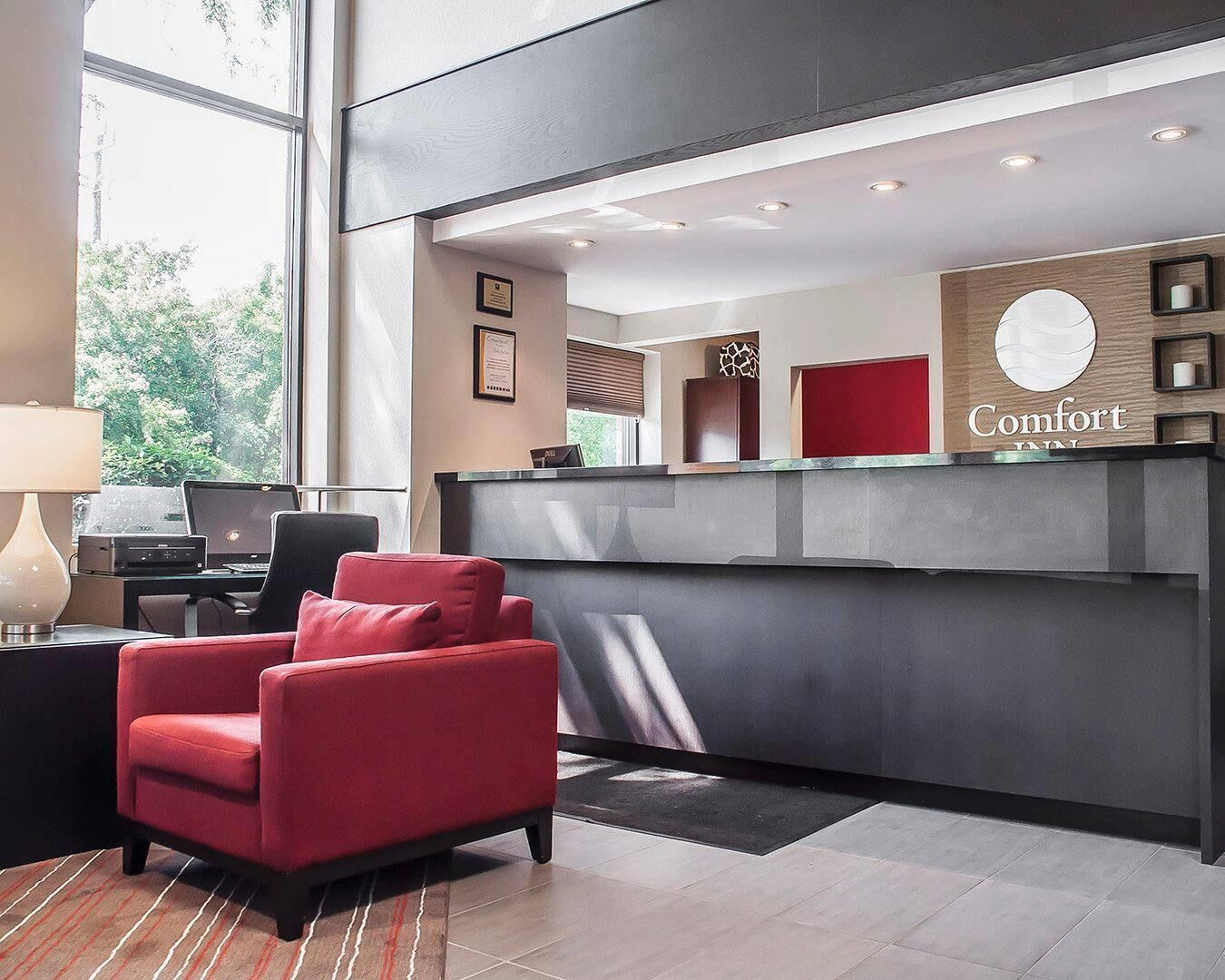 A hotel reception area with a modern design, featuring a reception desk, a red upholstered chair, and a laptop on a desk.