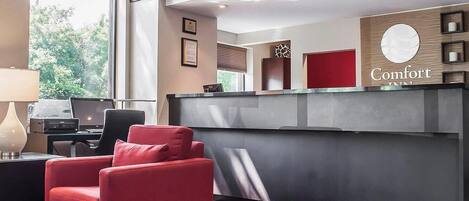 A hotel reception area with a modern design, featuring a reception desk, a red upholstered chair, and a laptop on a desk.