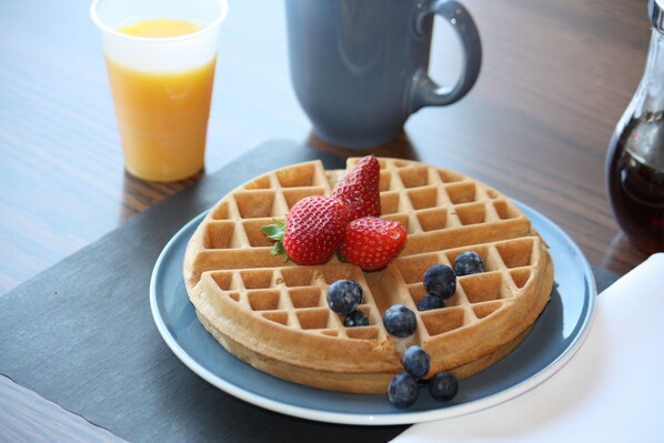 Een wafel met aardbeien en bosbessen op een bord, een glas sinaasappelsap en een karaf op de achtergrond.