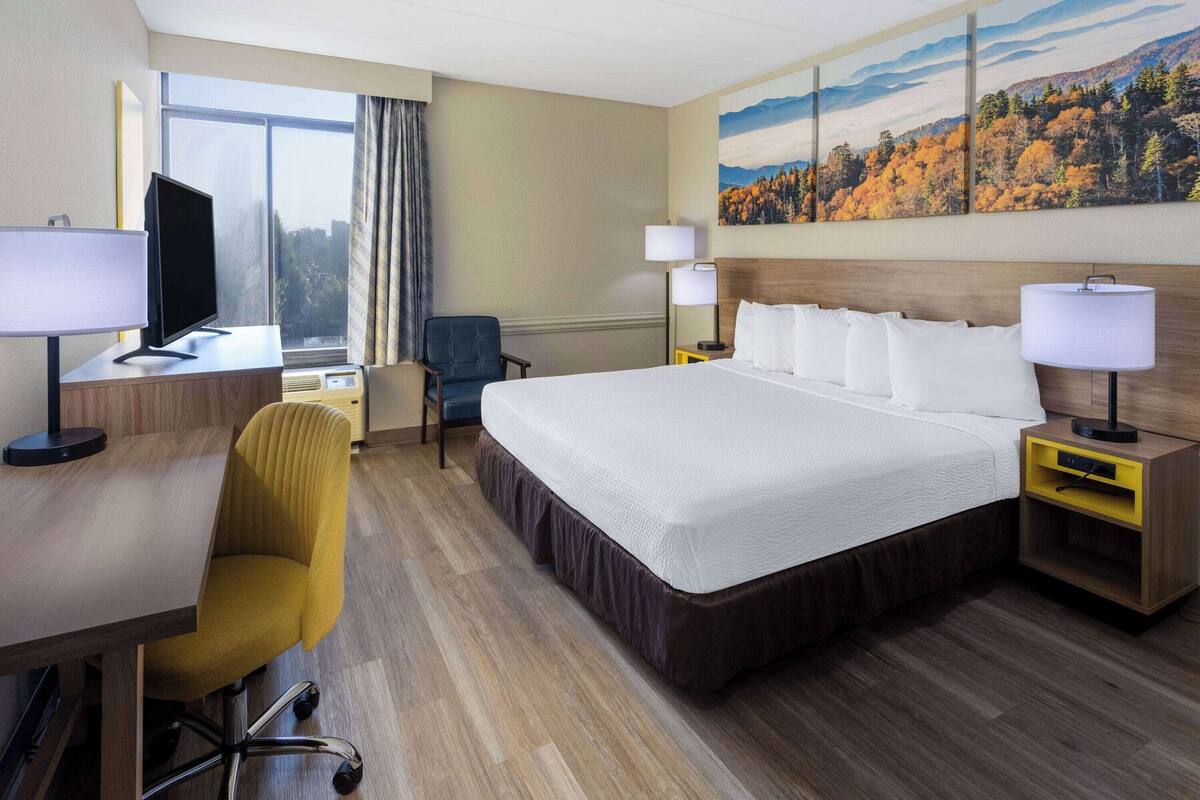 A hotel room with a large bed, a desk with a computer, a chair, two bedside tables with lamps, and a window with a view of trees.
