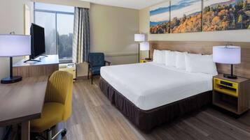 A hotel room with a large bed, a desk with a computer, a chair, two bedside tables with lamps, and a window with a view of trees.