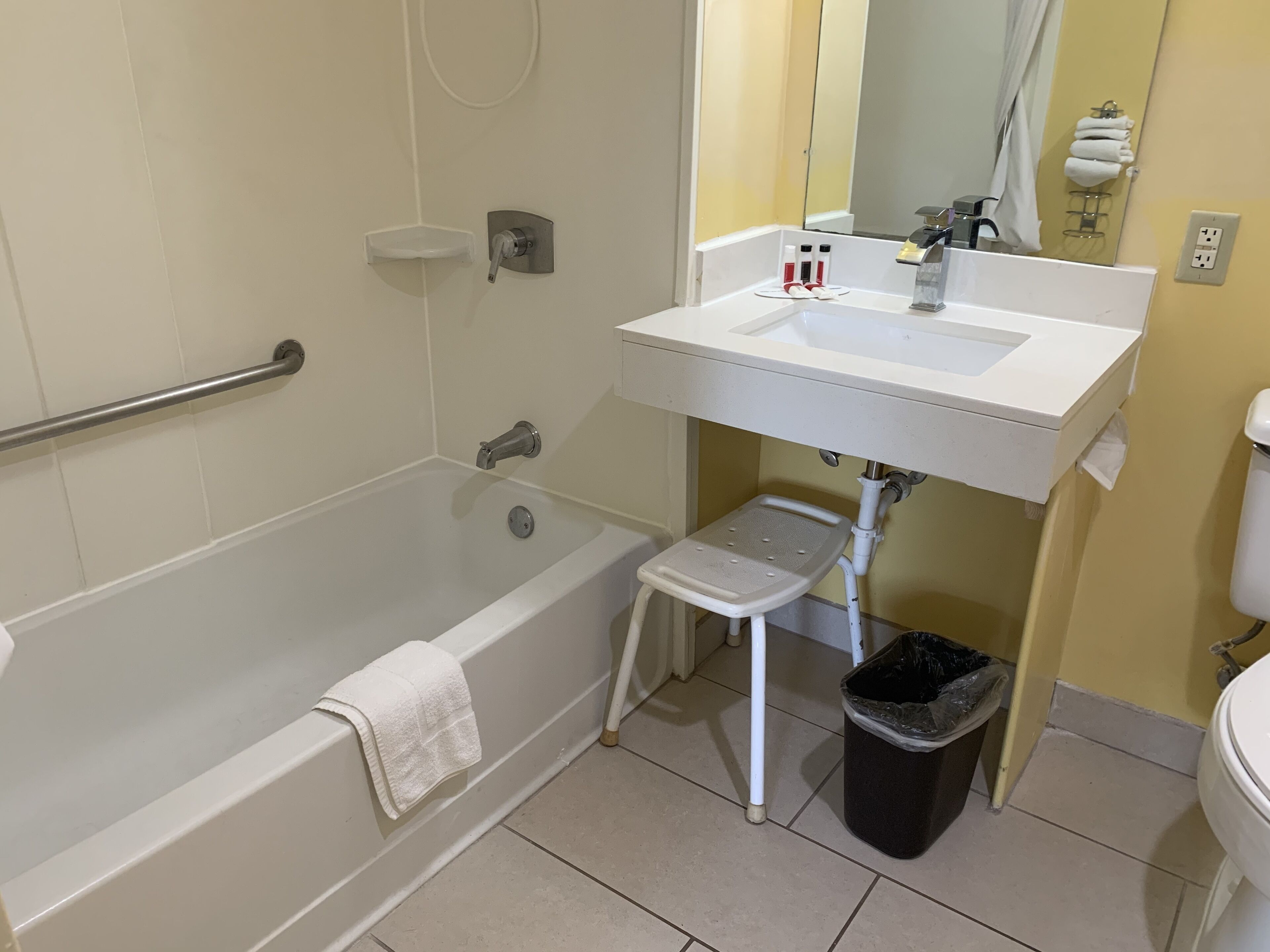 A bathroom with a bathtub, a white sink, a small stool, a trash bin, and a towel rack.
