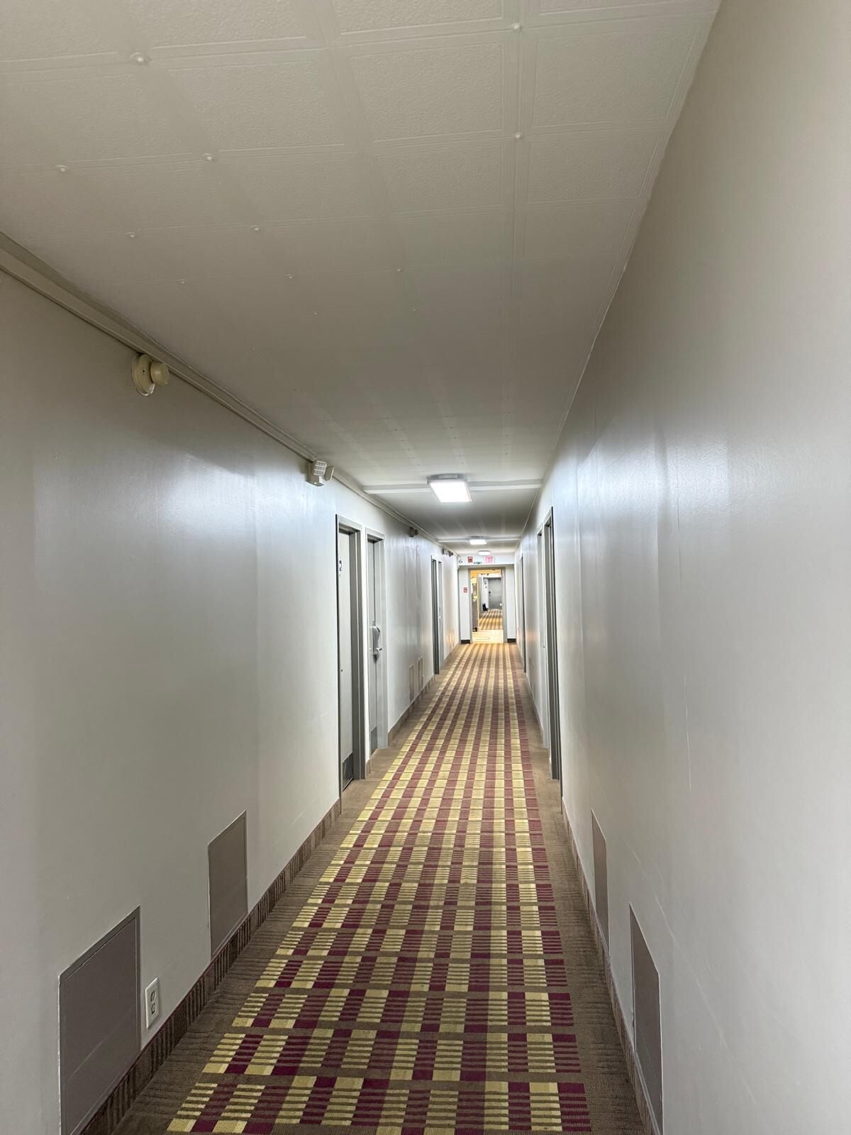 A long, narrow hotel corridor with patterned carpet, white walls, and doors on both sides.