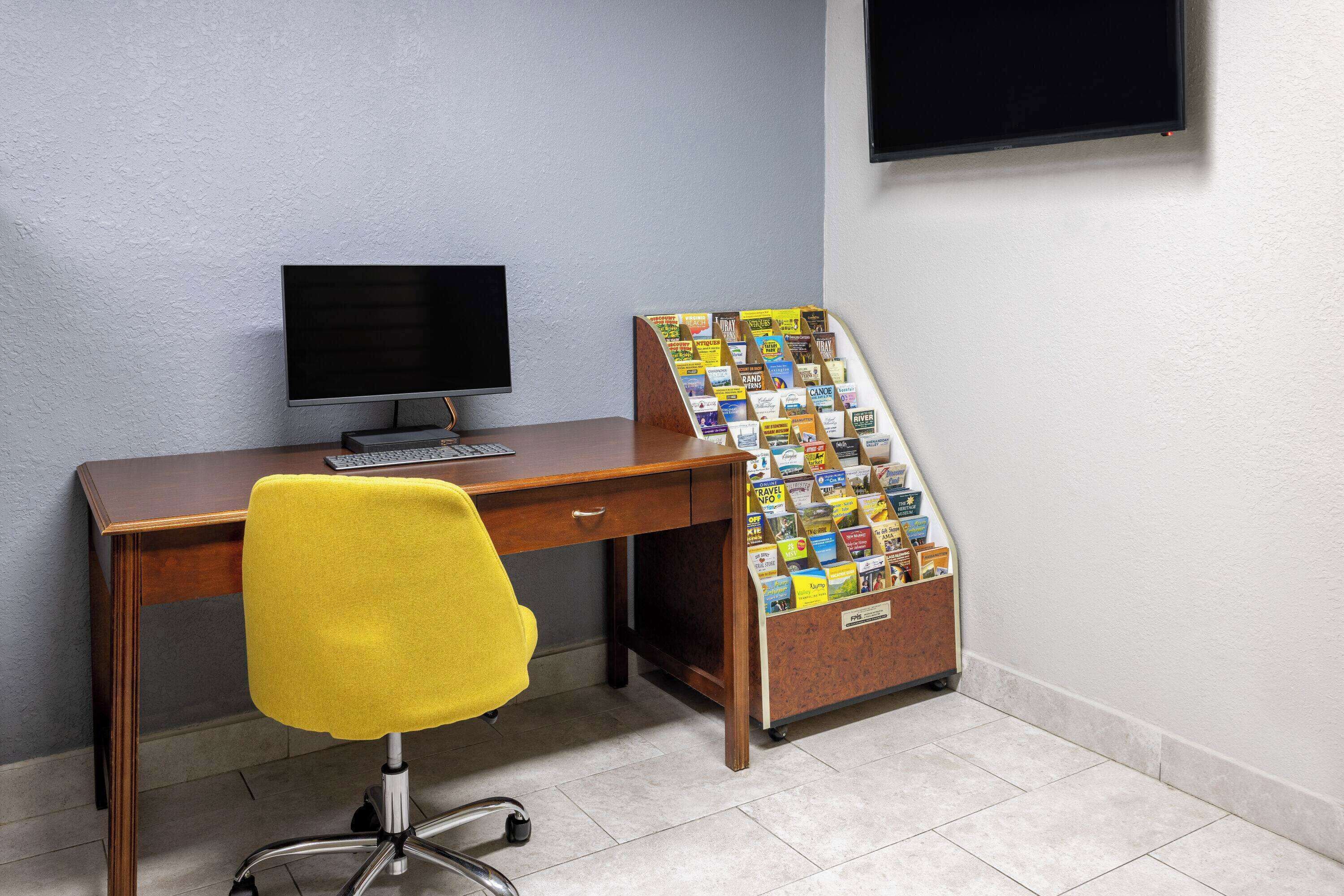Un escritorio de madera con un monitor de computadora, una silla de oficina amarilla y un tablón de anuncios con folletos de viajes.