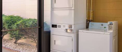 A laundry area with stacked washers and dryers, a window with a view of a tree and gravel path, and visible plumbing.