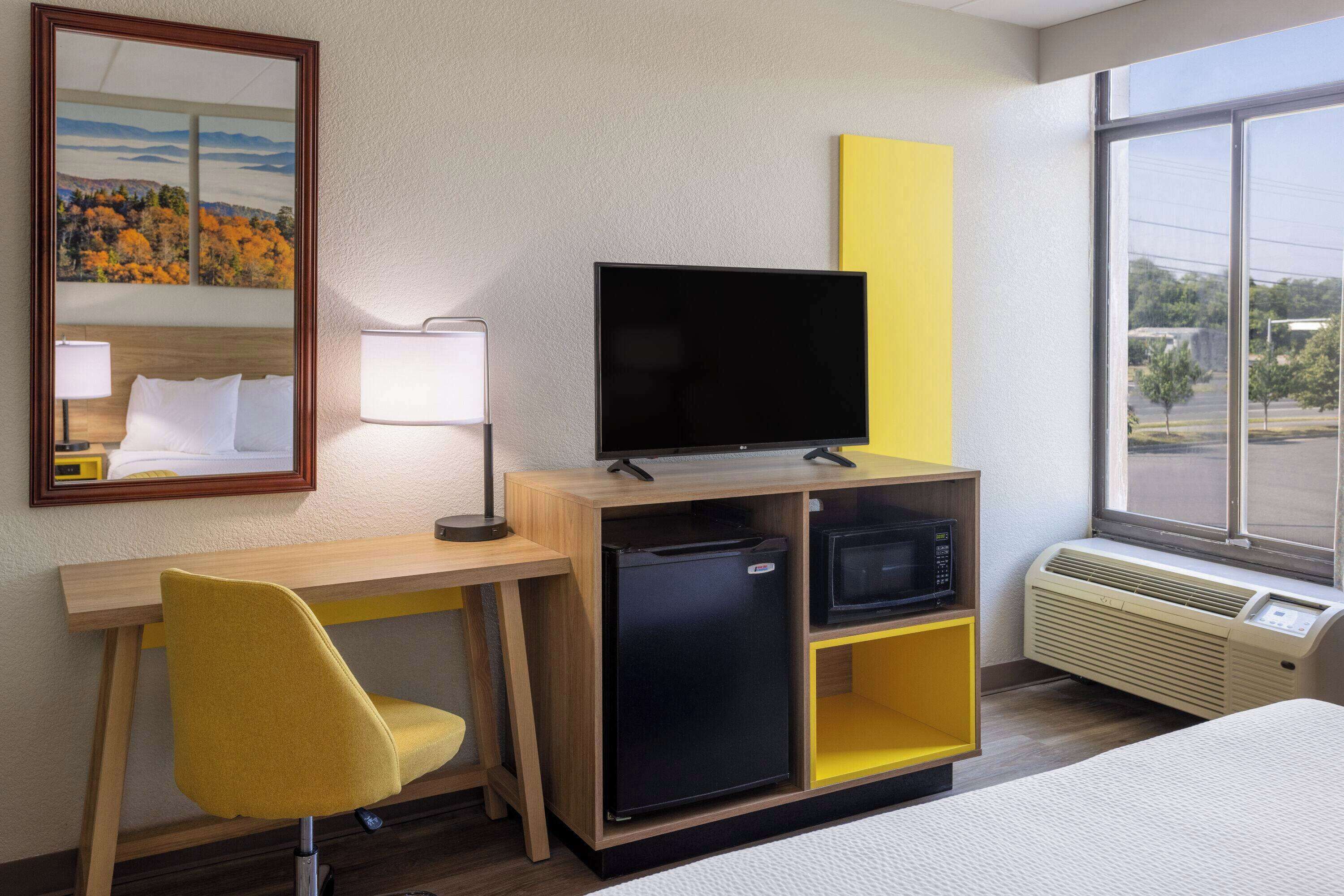 Une chambre d’hôtel équipée d’un lit, d’un bureau avec un ordinateur, d’un petit réfrigérateur, d’une télévision et d’une fenêtre donnant sur l’extérieur.