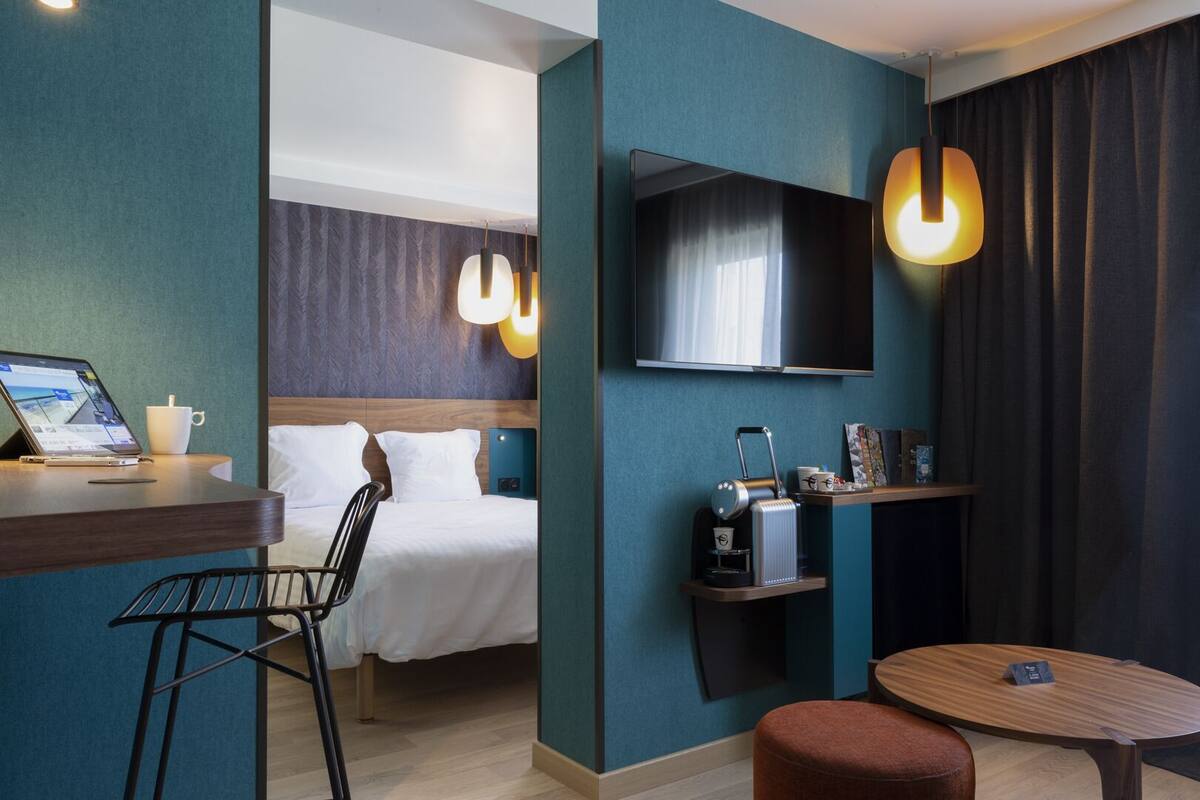 Habitación de hotel con cama, escritorio con computadora portátil, cafetera y televisor montado en la pared.
