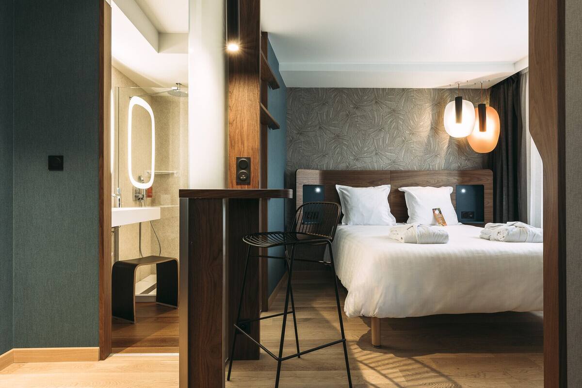 Una habitación de hotel moderna con una cama grande, una mesita de noche, una silla, un escritorio, un espejo y una lámpara fijada a la pared.