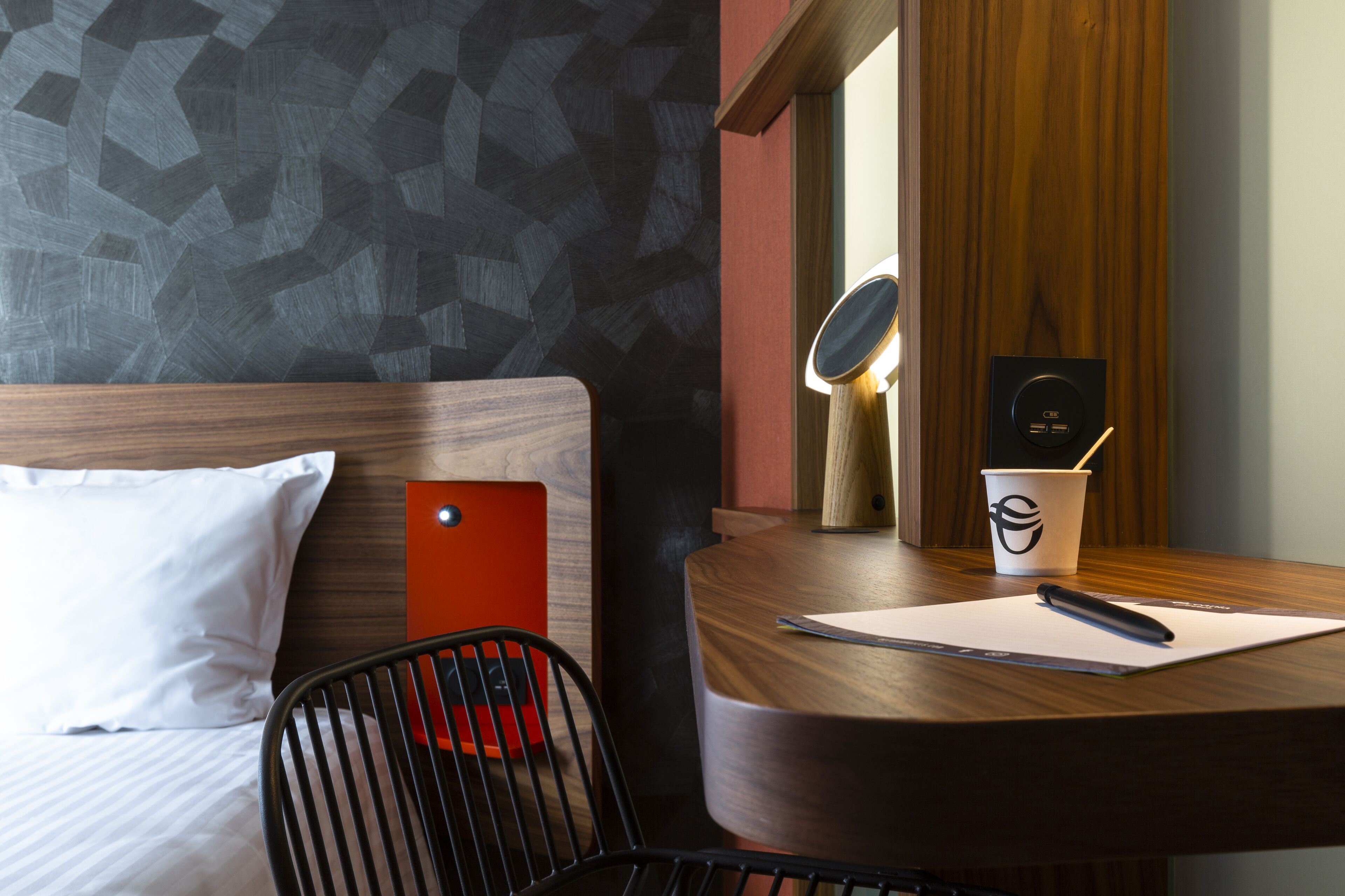 Une chambre d’hôtel avec un lit, un bureau avec une tasse, une chaise et un miroir fixé au mur.