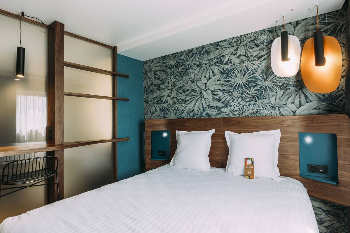 Habitación de hotel con cama, escritorio y silla. Hay luces fijadas a la pared y un panel decorativo.