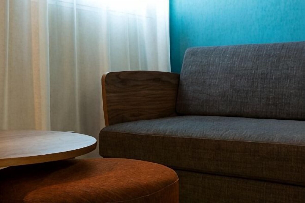 Ein Zimmer mit einer Couch, einem runden Holztisch und einem Fenster mit durchsichtigen Vorhängen.