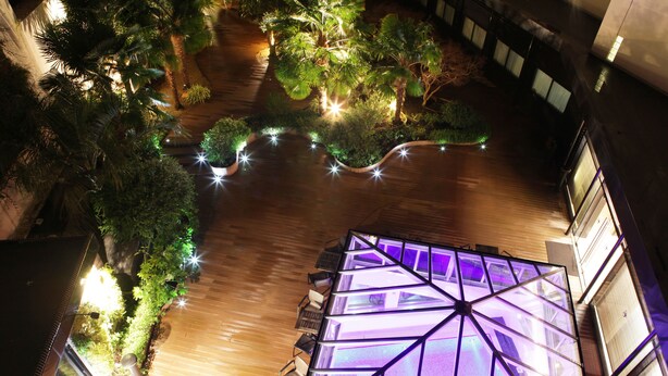 Vista aérea de un patio de hotel por la noche, con senderos iluminados, una fuente central y zonas verdes circundantes.