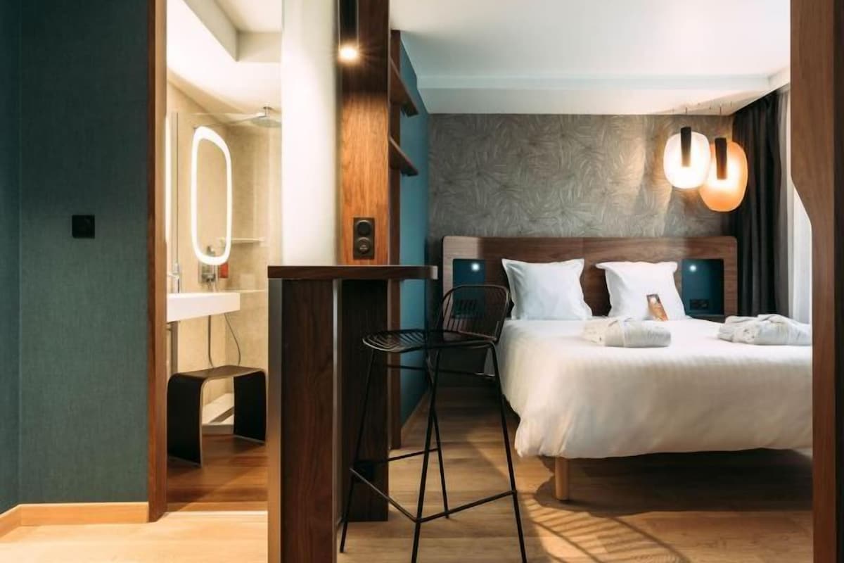 A modern hotel room with a wooden headboard, a large bed, a small stool, a vanity with a mirror, and a wall-mounted light fixture.