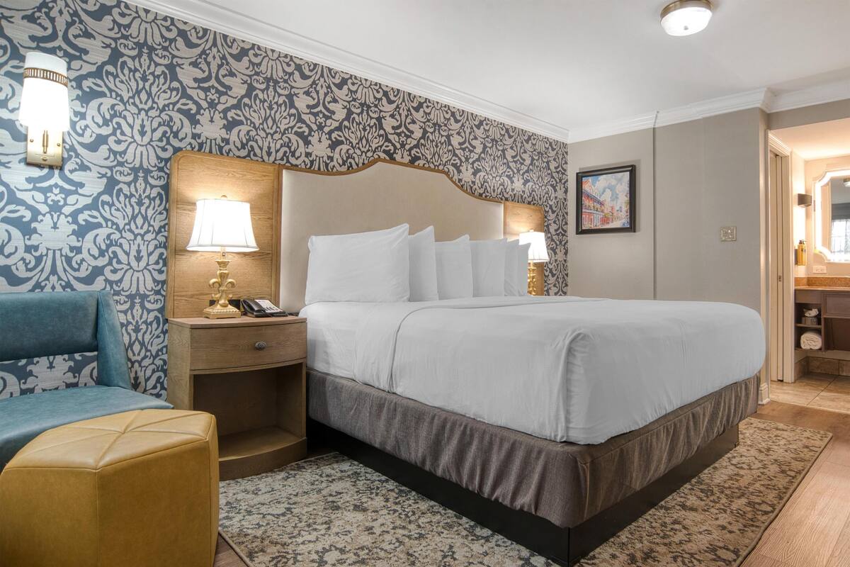 A hotel room with a large bed, a bedside table with a lamp, a nightstand, a chair, and patterned wallpaper.