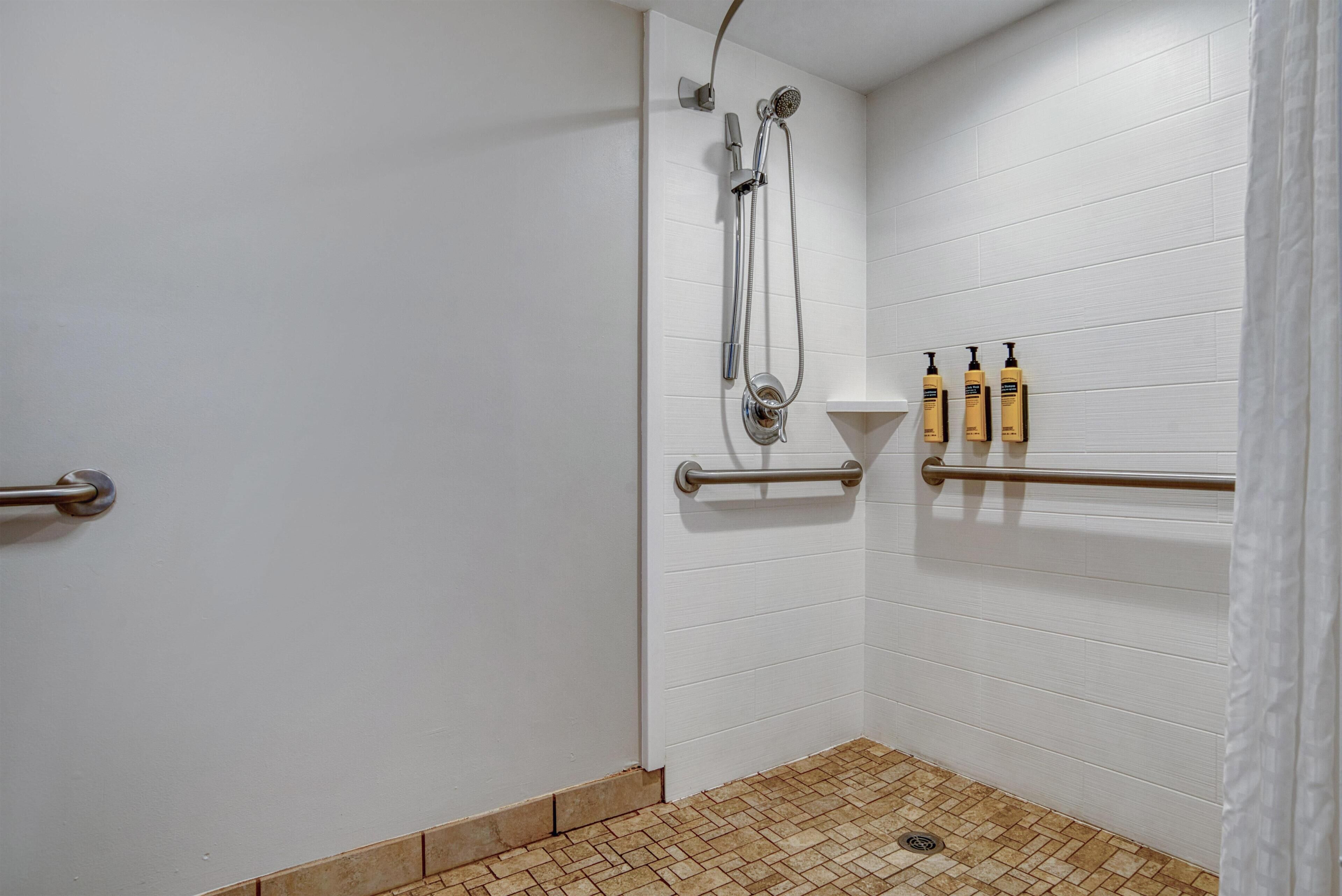 Un espace douche équipé d’un pommeau de douche à main, d’un robinet mural et d’une barre d’appui. On y trouve trois bouteilles de shampoing et d’après-shampoing sur une étagère.