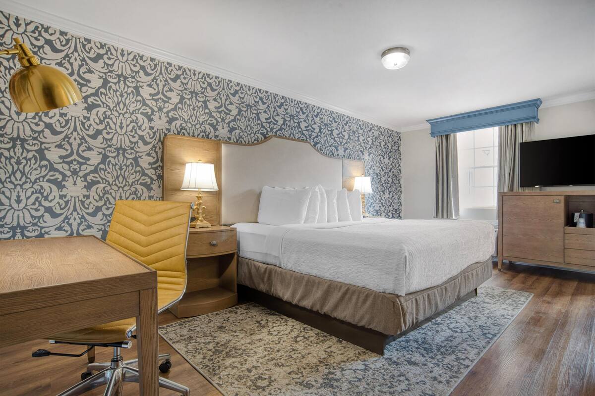 A hotel room with a bed, bedside lamps, a desk, a chair, a television, and patterned wallpaper.