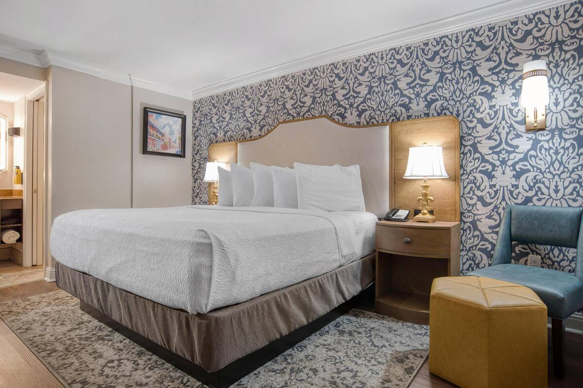 A neatly arranged hotel room with a large bed, a bedside table with a lamp, a chair, and a patterned wallpaper.