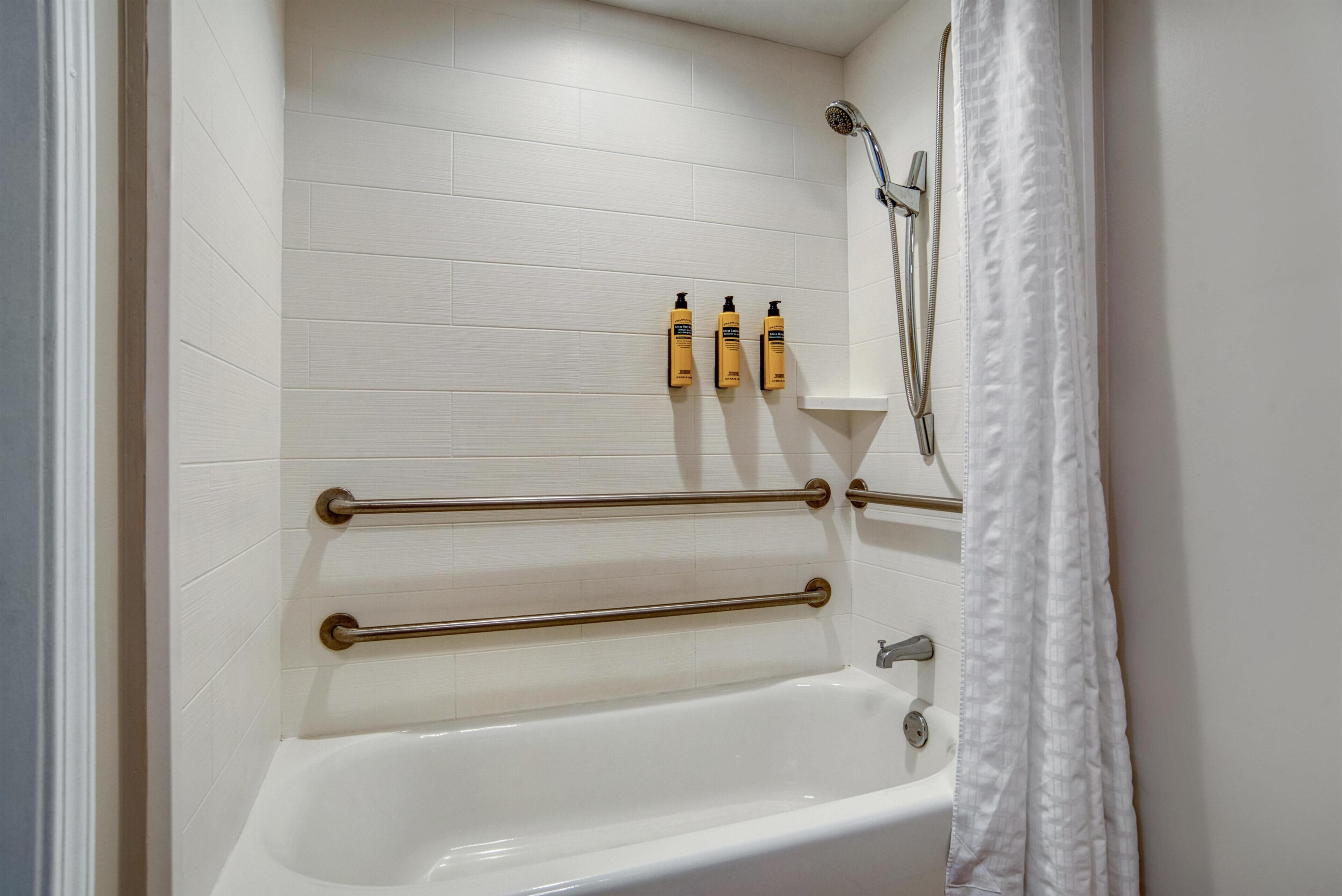 A bathroom with a white bathtub, grab bars, a showerhead, and three bottles of shampoo on the wall.