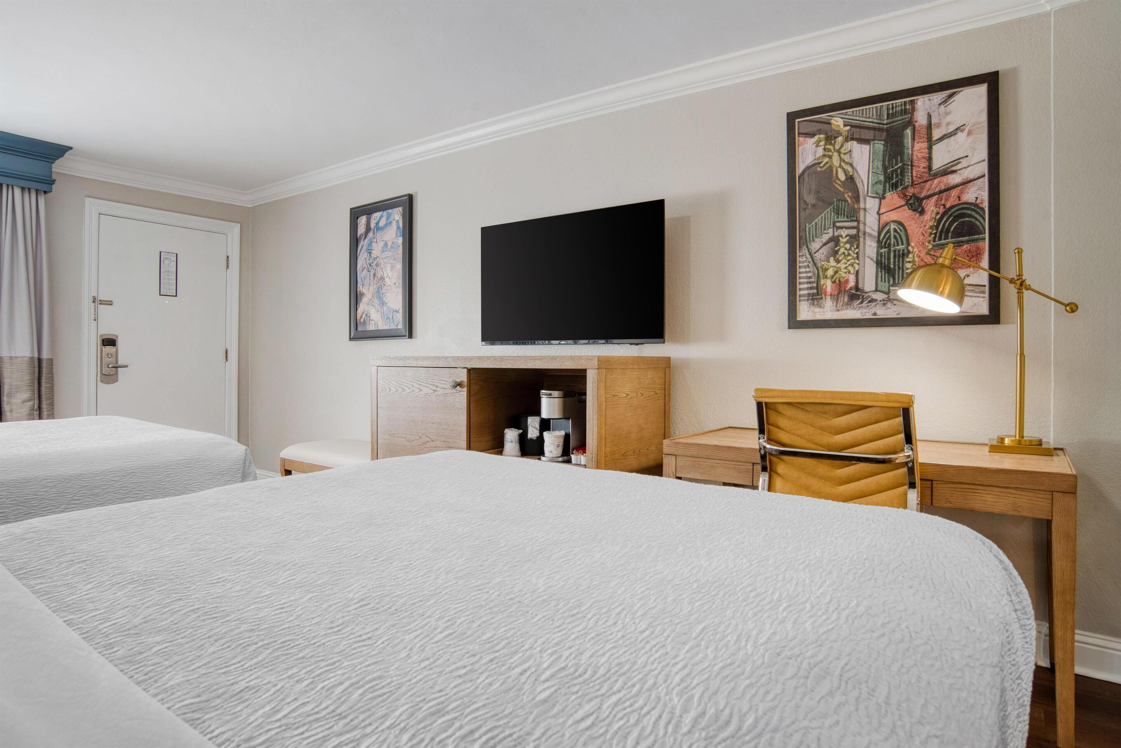 A hotel room with two beds, a flat-screen TV, a desk with a chair, and artwork on the walls.