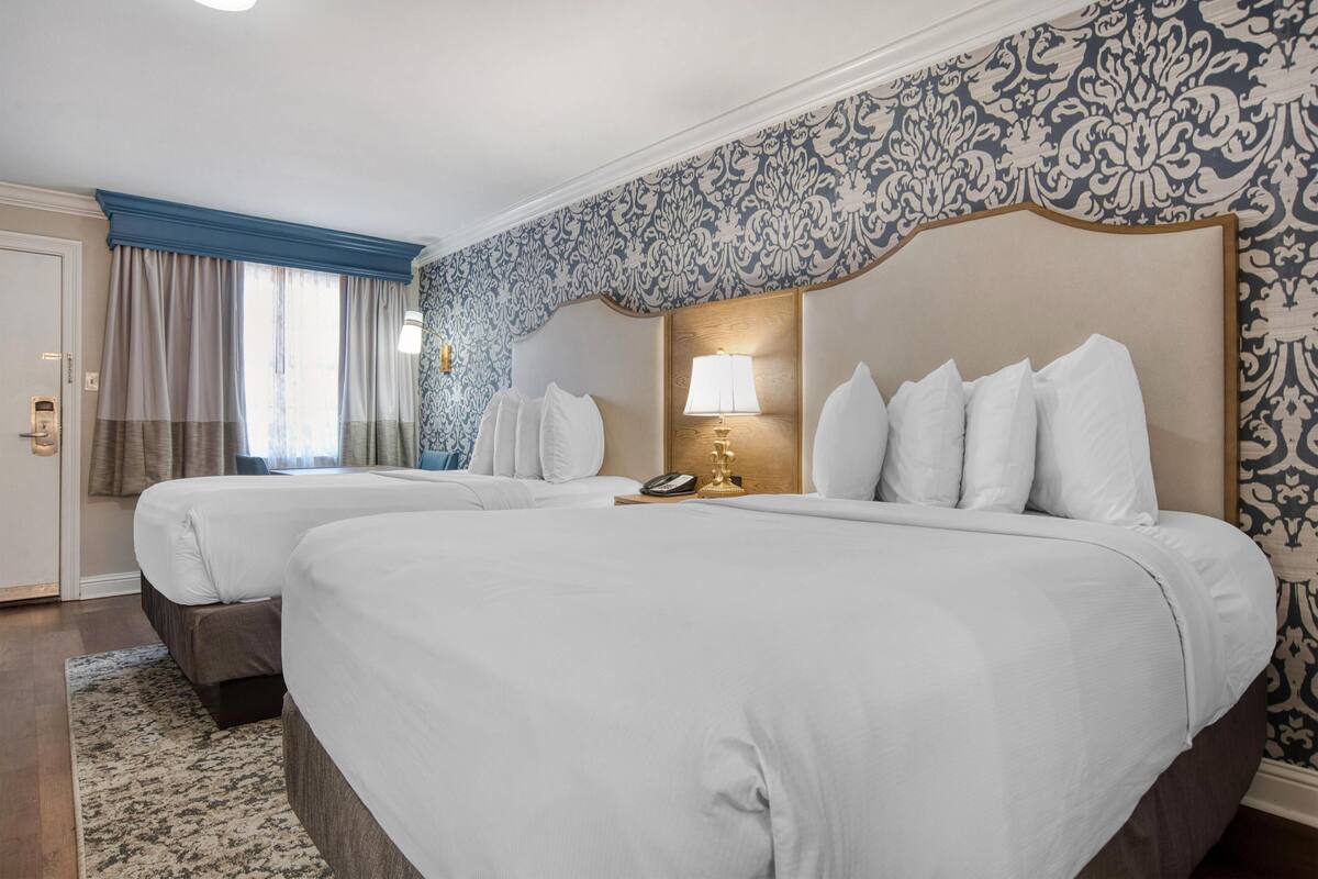 A hotel room with two beds, a nightstand with a lamp, and patterned wallpaper.
