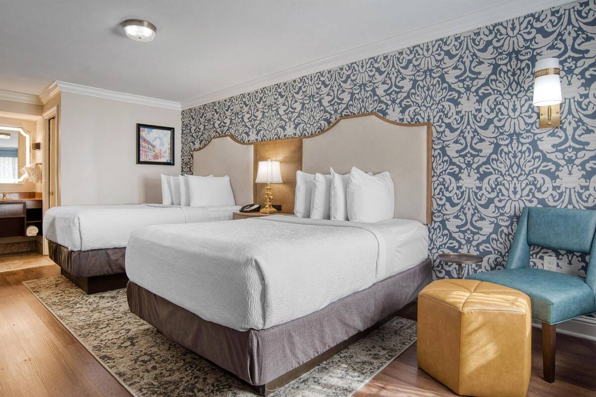 A hotel room with two beds, a blue patterned wall, a wooden floor, a blue armchair, and a small table with a lamp.