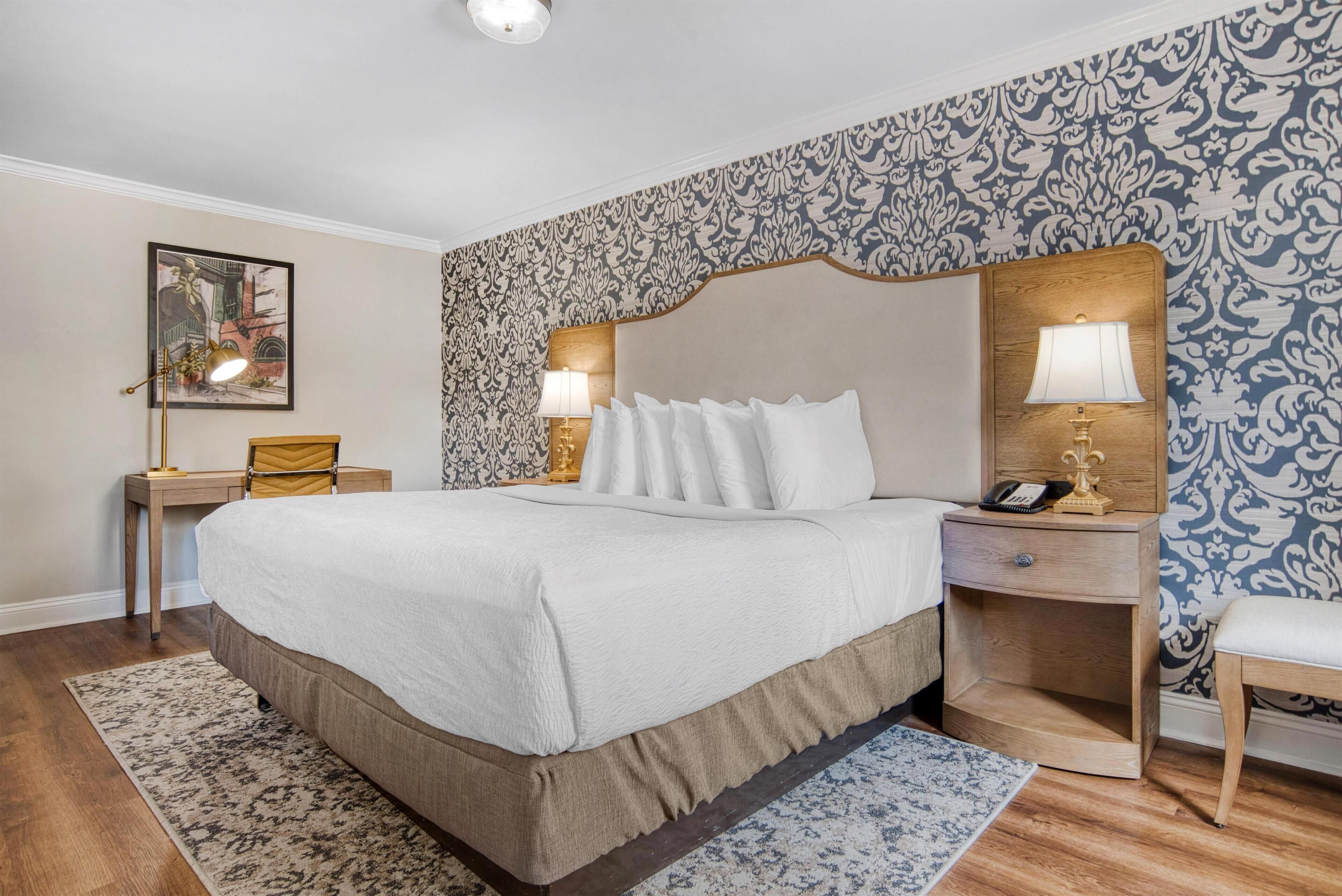 A neatly arranged bedroom with a large bed, two bedside tables, a nightstand, a chair, and a patterned wallpaper.