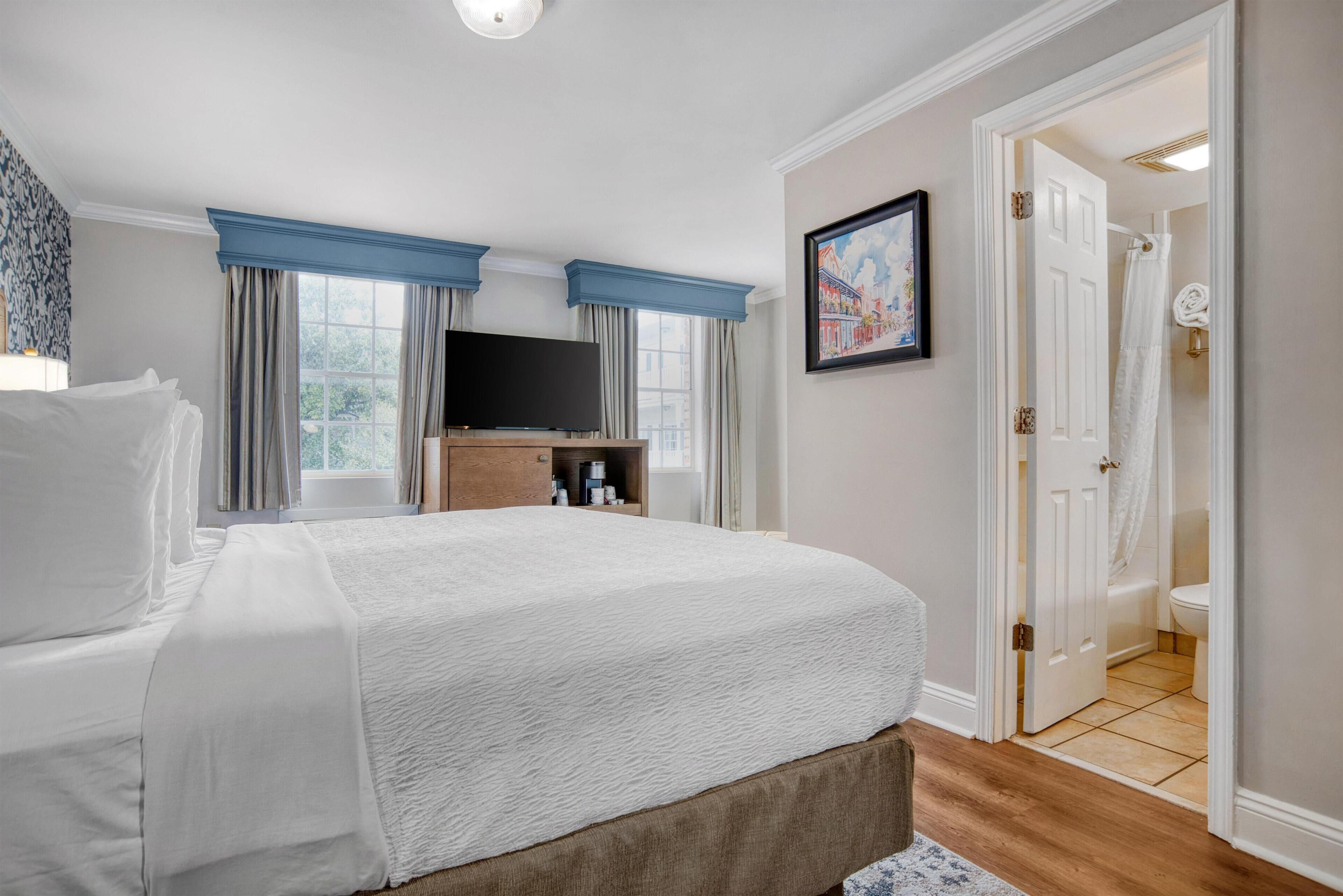 A bedroom with a large bed, a flat-screen TV, a wooden dresser, and a bathroom visible through an open door.