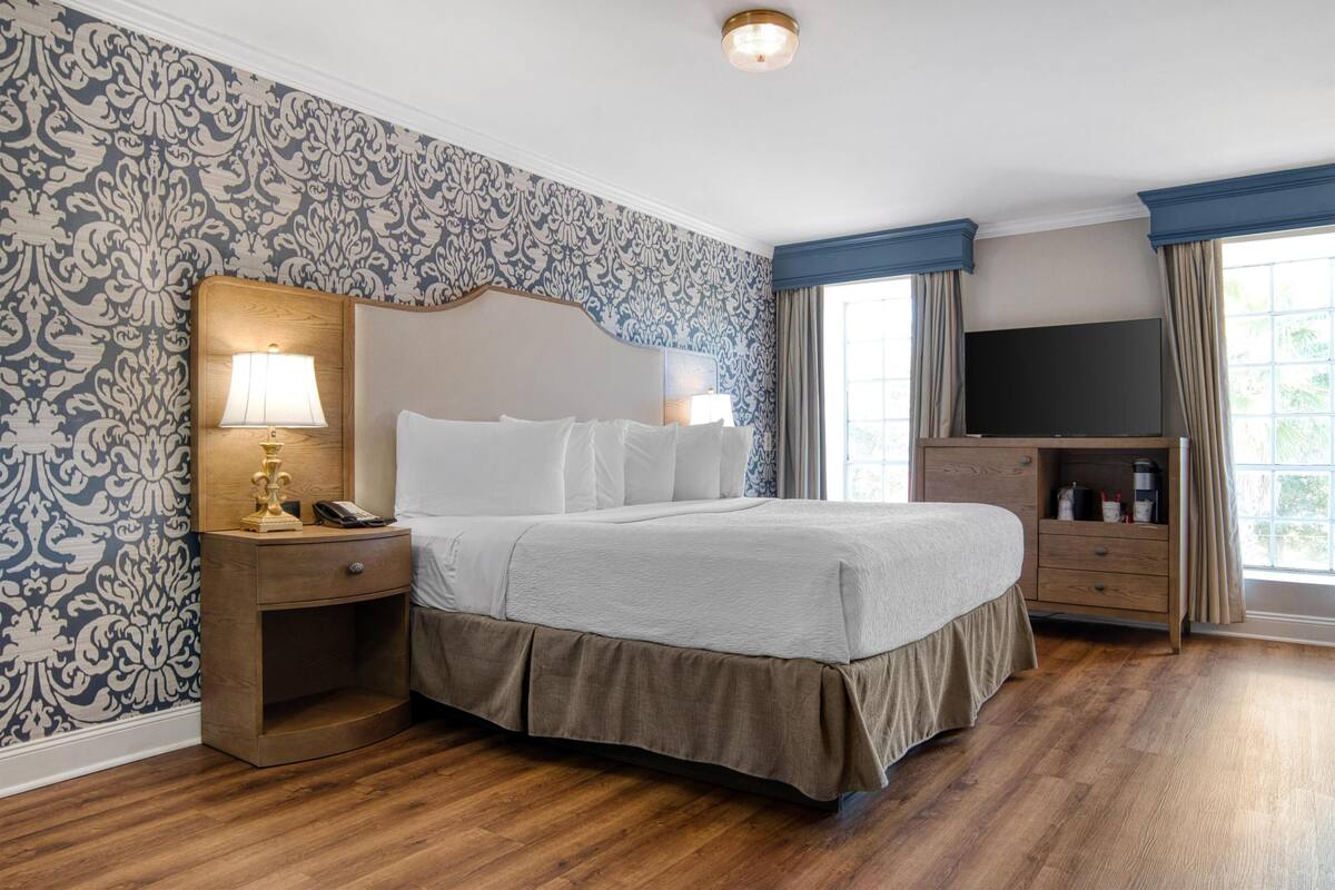 A hotel room with a large bed, a nightstand with a lamp, a dresser, a television, and a window with curtains.