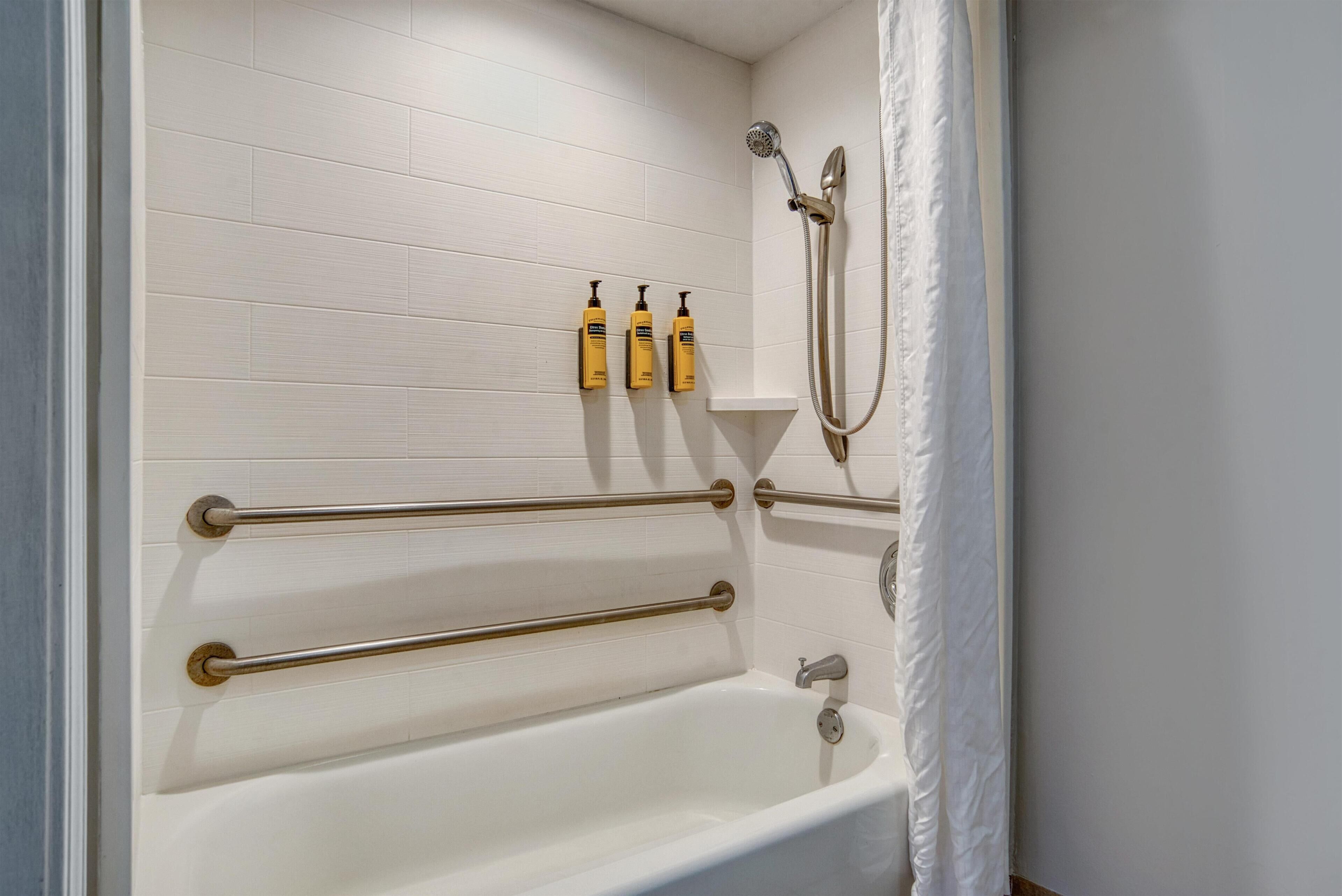 Une salle de bain avec une baignoire blanche, des barres d’appui, une pomme de douche et trois bouteilles de shampoing.