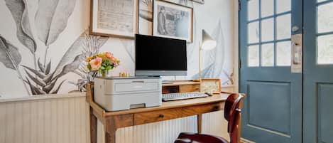 Ett skrivbord i trä med en skrivare, en datorskärm och ett tangentbord. Ovanför skrivbordet hänger en inramad tavla och framför det står en stol.