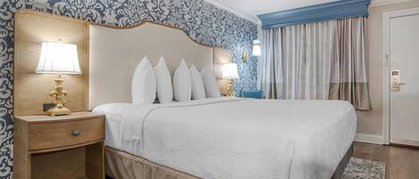 A neatly made bed with a white comforter and pillows, a wooden nightstand with a lamp, and a patterned wallpaper.