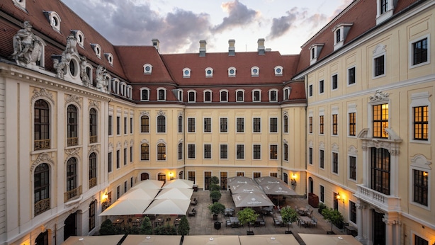 A courtyard with multiple tables and umbrellas, surrounded by classical architecture with ornate details and a red-tiled roof.