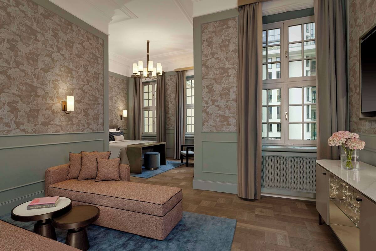 Ein Hotelzimmer mit Sofa, rundem Tisch, Schreibtisch und einem großen Fenster mit Vorhängen.