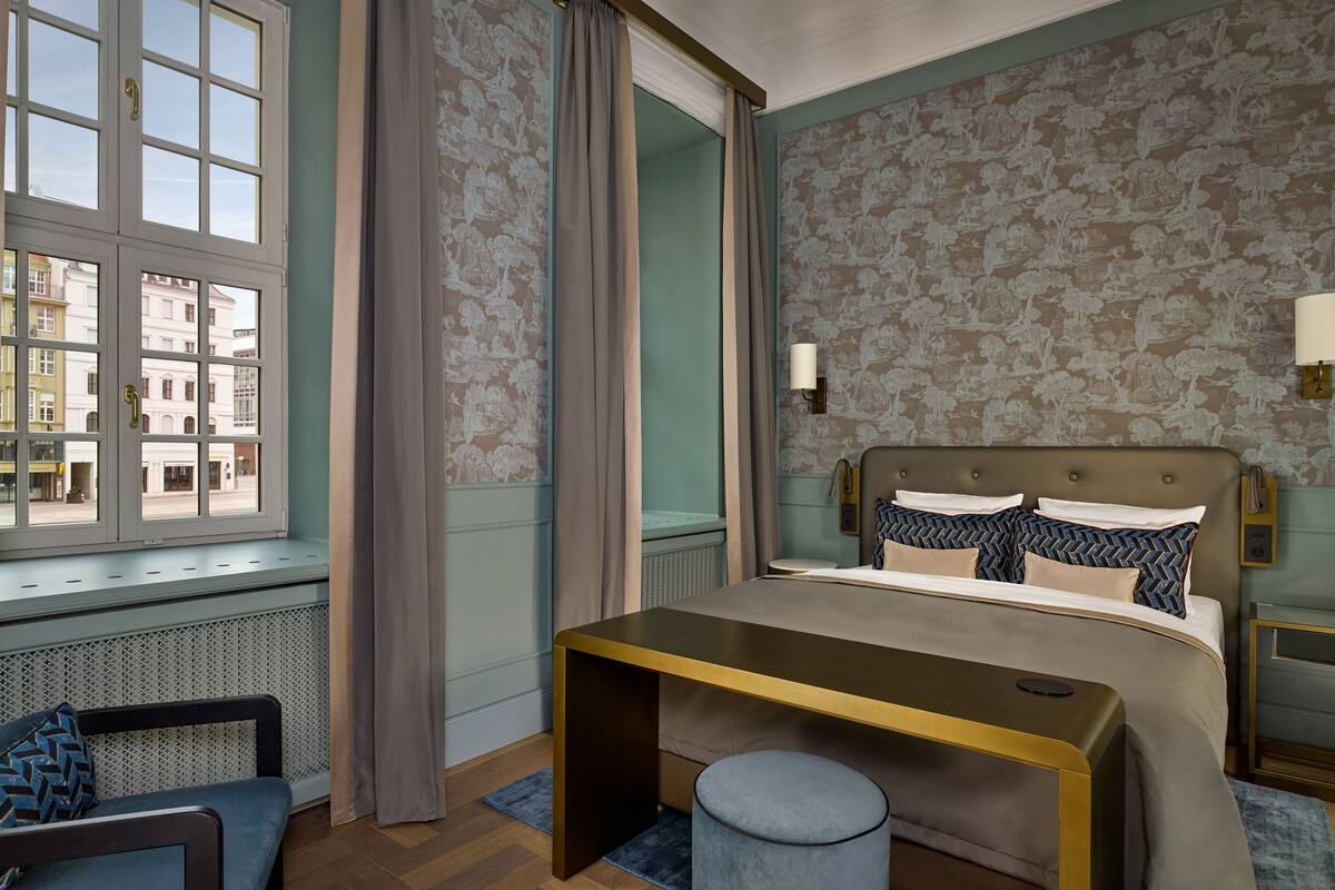 Ein Schlafzimmer mit Bett, Nachttisch, Stuhl, Fenster mit Vorhängen und Wand mit Blumenmuster.