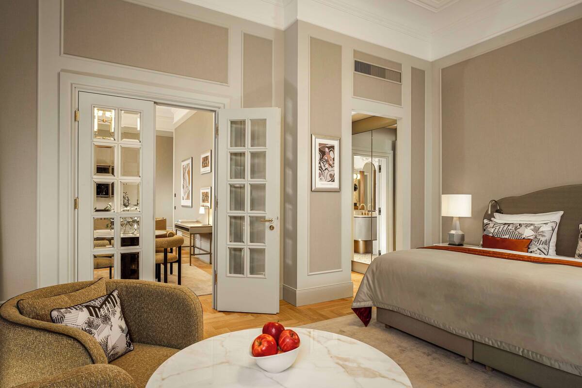 Ein Hotelzimmer mit einem Bett, einer Sitzecke mit einer Obstschale und einem Essbereich, der durch eine geöffnete Tür sichtbar ist.