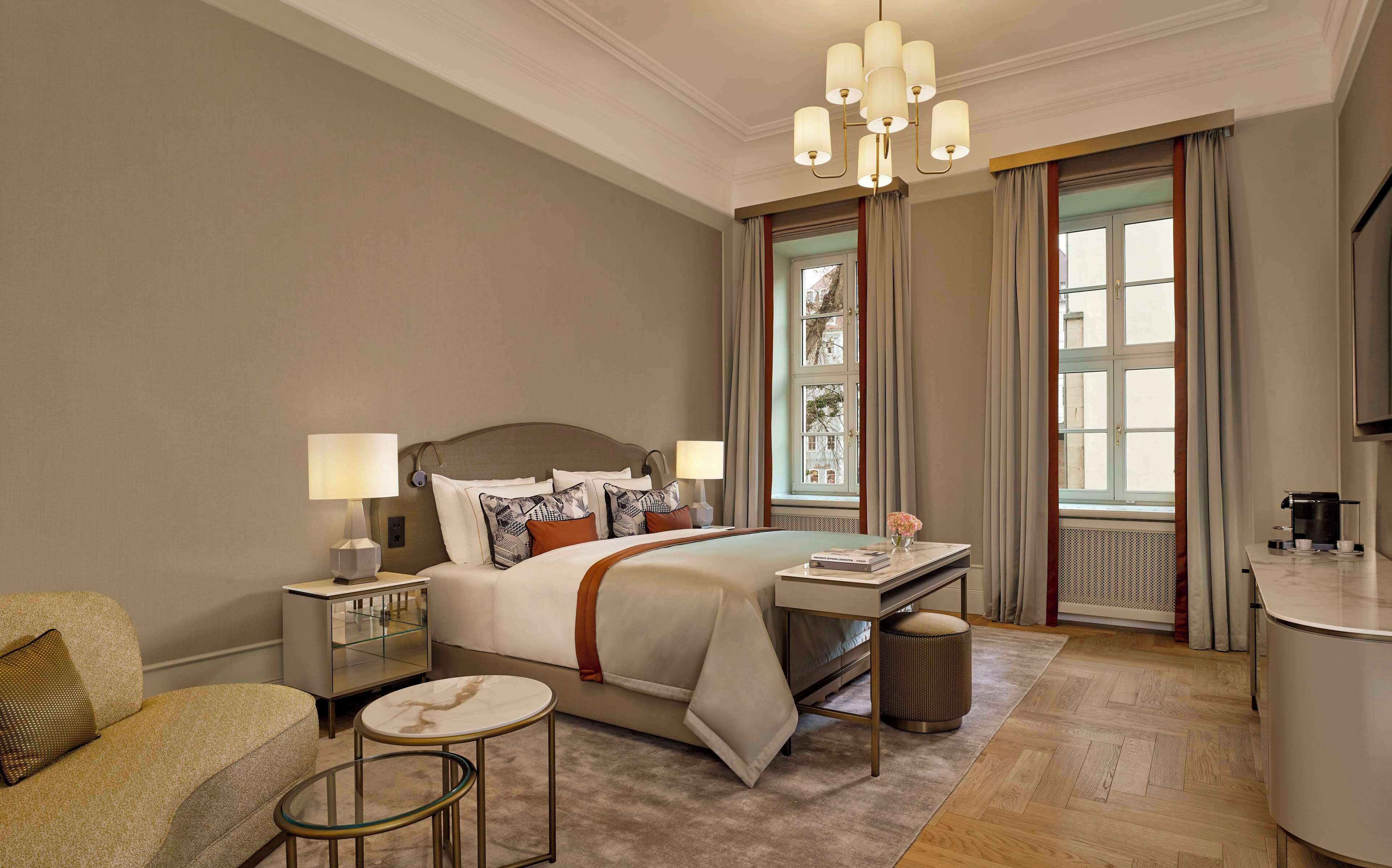 A hotel room with a large bed, bedside tables with lamps, a small round table, a sofa, a desk with a coffee maker, and a view of the city through large windows.